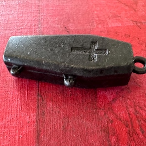 May include: A small black metal coffin-shaped pendant with a cross engraved on the top. The pendant has a loop at the top for attaching to a chain.