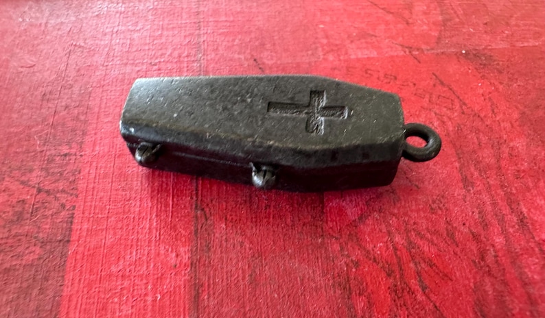 May include: A small black metal coffin-shaped pendant with a cross engraved on the top. The pendant has a loop at the top for attaching to a chain.