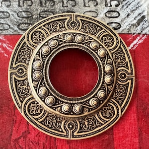 May include: A gold-toned metal decorative circle with intricate floral designs and a central ring. The circle has a raised, textured surface and a smooth, polished finish.