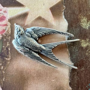 Może przedstawiać: Szczegółowa srebrna broszka w kształcie jaskółki z rozłożonymi skrzydłami. Pióra ptaka są misternie szczegółowe, a broszka ma teksturowany wygląd. Jaskółka jest symbolem wolności i nadziei.