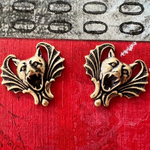 May include: Two gold-toned metal charms in the shape of a bat with a snarling face. The charms have a detailed design and are perfect for adding a gothic touch to jewelry or crafts.