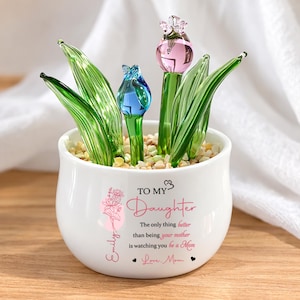 May include: A white ceramic pot with a floral design and the text "TO MY DAUGHTER". It contains glass tulip flowers in pink and blue, with green glass leaves. The pot is on a wooden surface.