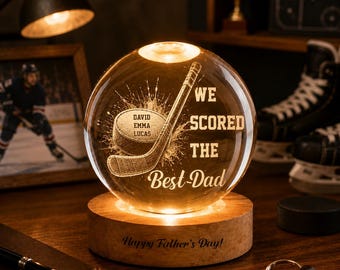 Op de afbeelding: Een verlichte glazen bol met een hockeyontwerp, met een hockeystick en puck, en de tekst "We Scored The Best-Dad". Hij staat op een houten voet met de inscriptie "Happy Father's Day!". Een ingelijste foto en hockeyschaatsen staan op de achtergrond.