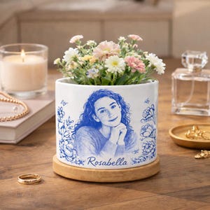 Op de afbeelding: Een witte keramische bloempot met een blauw portret en bloemenontwerp, met een verscheidenheid aan kleurrijke bloemen. De pot staat op een houten voet. De naam "Rosabella" is afgedrukt.