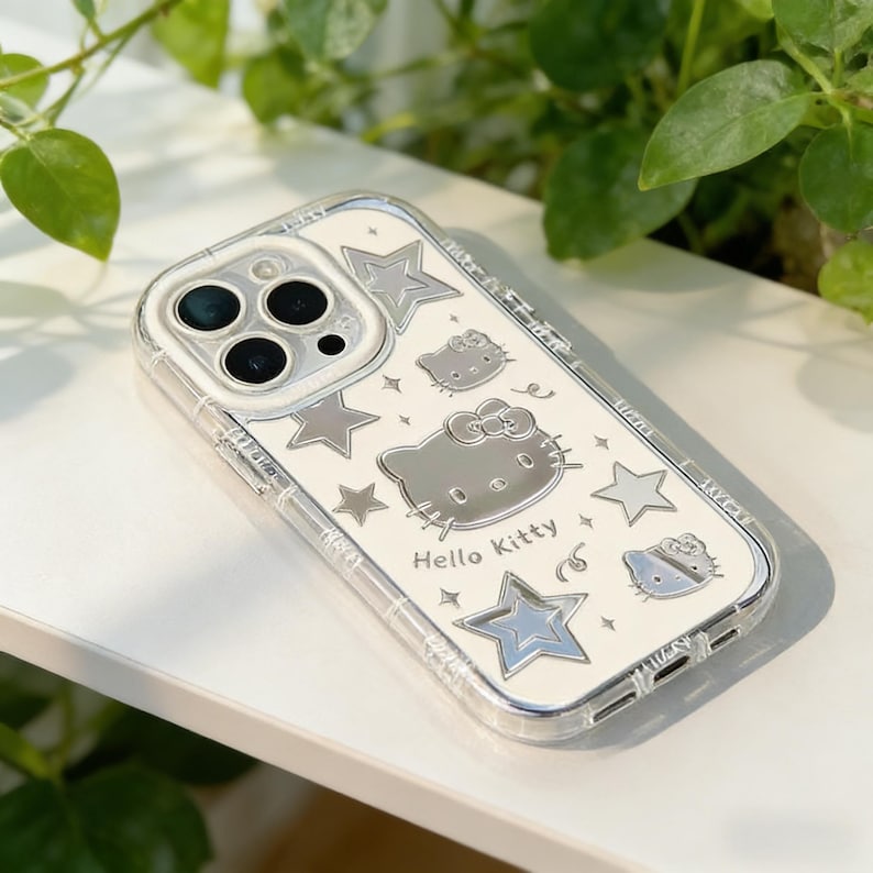 Może przedstawiać: Przezroczyste etui na telefon z srebrnym wzorem Hello Kitty. Etui ma białe tło ze srebrnymi gwiazdami i ilustracjami twarzy Hello Kitty. Tekst "Hello Kitty" jest wydrukowany na dole.