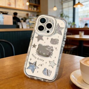 Może przedstawiać: Przezroczyste etui na telefon z motywem Hello Kitty. Etui posiada srebrną grafikę Hello Kitty, gwiazdki i napis "Hello Kitty". Etui leży na drewnianym stole.