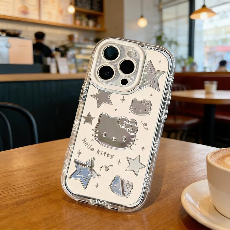 Może przedstawiać: Przezroczyste etui na telefon z motywem Hello Kitty. Etui posiada srebrną grafikę Hello Kitty, gwiazdki i napis "Hello Kitty". Etui leży na drewnianym stole.