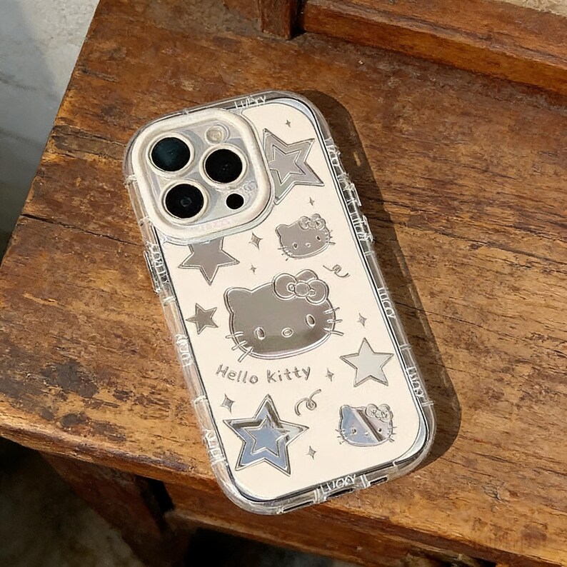 Może przedstawiać: Przezroczyste etui na telefon z srebrnym lustrzanym wnętrzem z motywem Hello Kitty i gwiazd. Etui ma przezroczystą obw&oacute;dkę, a na nim nadrukowano napis "Hello Kitty".