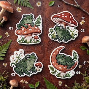 May include: Four embroidered patches featuring green frogs. Patch 01 shows a frog on a mushroom. Patch 02 features a frog under a large mushroom. Patch 03 depicts a frog with white flowers. Patch 04 shows a frog resting on a crescent moon shape.
