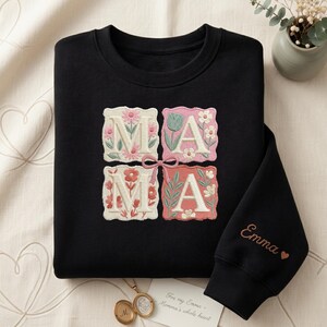 May include: Black sweatshirt with embroidered floral "MAMA" design. Each letter is in a square with a different floral pattern. The sleeve has the name "Emma" embroidered in a copper color with a heart symbol.