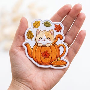 May include: An embroidered patch featuring a cream and orange kitten sitting inside an orange pumpkin-shaped teapot. The design includes small embroidered autumn leaves in yellow, green, and red colors surrounding the kitten and teapot.