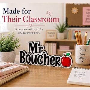 May include: A black and white custom name sign reading Mr. BOUCHER with a red apple graphic on the right side. The sign is made of thick material with a white outline and sits on a desk alongside a potted plant, a pencil holder, and notebooks.