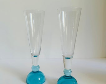 Puede incluir: Dos copas de champán de vidrio transparente con bases de vidrio azul. Las bases son redondas y tienen un pequeño pomo en la parte inferior.