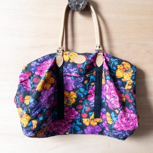 May include: A floral print travel bag with brown leather handles and black straps. The bag is hanging on a hook.