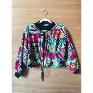 Puede incluir: Una chaqueta bomber corta vintage con un diseño de patchwork vibrante. La chaqueta presenta una mezcla de estampados florales, de lunares y a rayas en rosa, verde, negro y blanco. Tiene cuello, puños negros y un dobladillo con cordón. La chaqueta está colgada en una percha blanca.