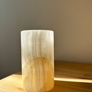 Puede incluir: Un jarrón cilíndrico de alabastro blanco y beige con una superficie lisa y pulida.