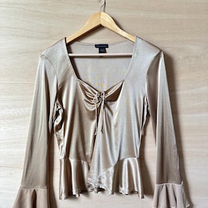 May include: A long-sleeved, gold satin blouse with a ruffled neckline and bell sleeves. The blouse has a tie at the neckline and a ruffled hem.