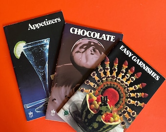 May include: Three cookbooks with colorful covers. The first cookbook is titled "Appetizers" and features a martini glass with a lime wedge. The second cookbook is titled "Chocolate" and features a chocolate dessert. The third cookbook is titled "Easy Garnishes" and features a watermelon garnished with fruit and candy.