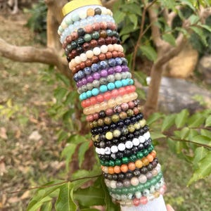 May include: A stack of beaded bracelets in various colors, including green, white, orange, black, and purple. The bracelets are made of different types of stones and are displayed on a white cylindrical stand against a blurred background of greenery and tree branches.