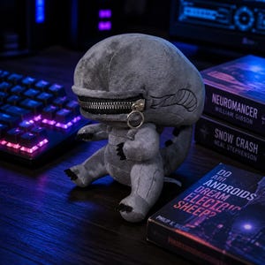 Puede incluir: Un peluche gris con forma de alienígena, con una boca con cremallera y un pequeño anillo metálico. El juguete está sobre una superficie de madera, junto a libros con títulos como "Neuromancer" y "Do Androids Dream of Electric Sheep?"