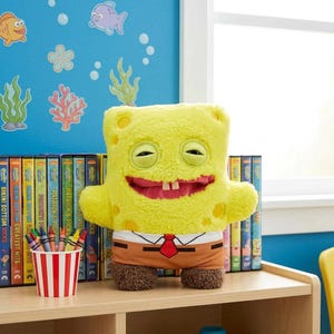 Könnte beinhalten: Ein gelbes SpongeBob Schwammkopf Plüschtier mit rotem Mund, zwei Vorderzähnen und braunen Shorts. Das Spielzeug steht auf einem Holzregal mit bunten DVDs und einem Stifthalter.