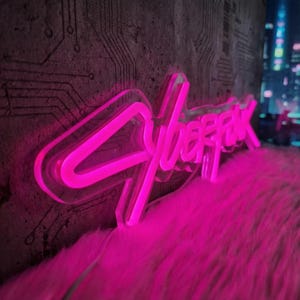 May include: A neon sign displaying the word "Cyberpunk" in vibrant pink. The sign is set against a textured grey wall with circuit board designs. It rests on a fluffy pink surface.