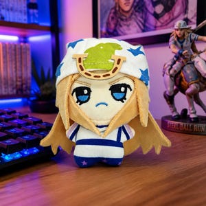 May include: A plush doll featuring blonde hair, a white hat with blue stars and a green patch, and a tan horseshoe-shaped accessory on the forehead. The doll has embroidered blue eyes, a small blue mouth, and a white body with blue horizontal stripes.