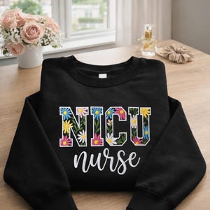 May include: Black crewneck sweatshirt with the words "NICU nurse" in white script. The letters "NICU" are filled with colorful floral designs, including daisies and other flowers in shades of pink, yellow, and blue.