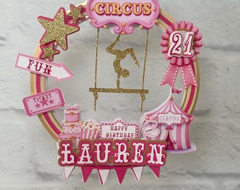 May include: A pink and gold circus-themed cake topper with the name "Lauren" and the number "21". The design includes a circus tent, stars, and the word "Circus". The topper is decorated with glitter and pink accents.