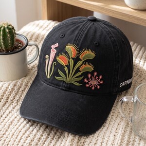 May include: A black baseball cap featuring an embroidered design of various carnivorous plants, including a pink pitcher plant, green and orange Venus flytraps, and a small pink sundew. The word CARNIVOROUS is partially visible on the side panel.