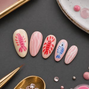 May include: A set of five painted fingernails. The nails feature designs of a pink lobster, a red lobster, a blue fish, and pink and white wavy stripes. The nails are on a dark surface with a gold brush and a small dish of pink paint.