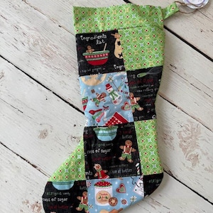 May include: A green, blue, and black Christmas stocking with a patchwork design featuring gingerbread men, cookie cutters, and baking ingredients. The stocking has a green cuff with a white tag that says "Handmade with Love".