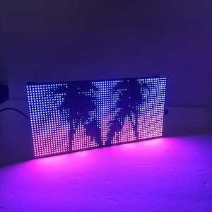 May include: A rectangular LED display with a black frame. The display shows a graphic of stylized mountain peaks, illuminated with a gradient of pink and blue lights. The display is plugged in.