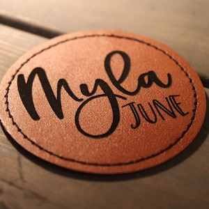 Pode incluir: Um patch redondo de couro castanho com o nome "Myla" em fonte cursiva e a palavra "JUNE" abaixo, ambos em preto. O patch tem uma borda costurada e está sobre um fundo de madeira.