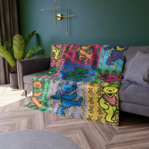 May include: A colorful throw blanket featuring a patchwork design with dancing bear graphics in various colors. The blanket is draped over a gray sofa, showcasing its vibrant hues of pink, blue, green, and yellow. The bears are in different poses.