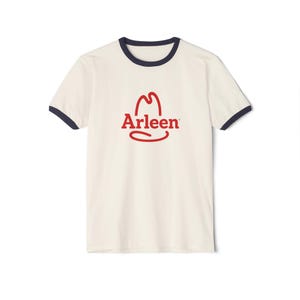 Puede incluir: Una camiseta tipo ringer de color crema con mangas cortas y cuello redondo y puños en azul marino oscuro. El centro del pecho presenta un logotipo gráfico rojo que consiste en el contorno de un sombrero de vaquero sobre el texto Arleen.