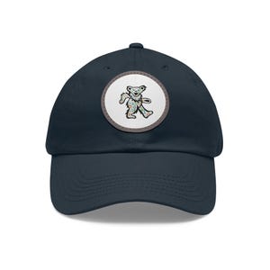 Puede incluir: Gorra de béisbol azul marino con un parche circular con un diseño de oso colorido. El oso tiene una pose de baile. El parche está bordeado por un anillo gris. La gorra tiene una visera curva y una corona estructurada.
