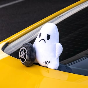 Könnte beinhalten: Ein weißes Plüschgespenst mit traurigem Gesicht und kleinen Flügeln, das ein graues Spielzeug-Autorad hält. Auf dem Gespenst ist die Zahl -10° aufgedruckt. Das Spielzeug befindet sich an der gelben Karosserie eines Autos.