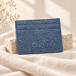 May include: A rectangular blue card holder featuring an embossed pattern of a koi fish swimming among lotus flowers and swirling water motifs. The accessory has a horizontal slot design with visible stitching along the edges and a flat surface.
