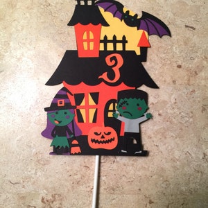 May include: A Halloween-themed cake topper featuring a black and orange haunted house with a witch, a Frankenstein, and a pumpkin. The house has a number "3" on the front.