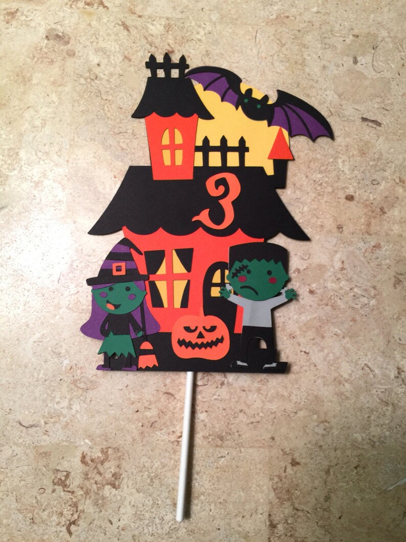 May include: A Halloween-themed cake topper featuring a black and orange haunted house with a witch, a Frankenstein, and a pumpkin. The house has a number "3" on the front.