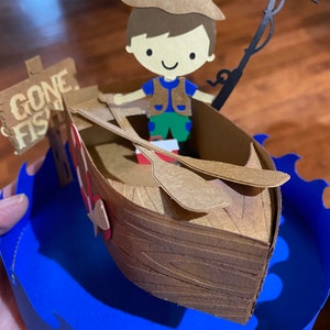 Puede incluir: Una figurita de papel de un ni&ntilde;o con un sombrero marr&oacute;n y un chaleco, sosteniendo una ca&ntilde;a de pescar y sentado en un bote de madera marr&oacute;n. El bote est&aacute; sobre una base de onda de papel azul con un letrero que dice "Gone Fishin".