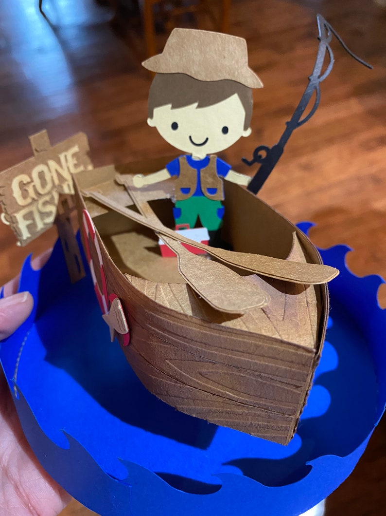 Puede incluir: Una figurita de papel de un ni&ntilde;o con un sombrero marr&oacute;n y un chaleco, sosteniendo una ca&ntilde;a de pescar y sentado en un bote de madera marr&oacute;n. El bote est&aacute; sobre una base de onda de papel azul con un letrero que dice "Gone Fishin".