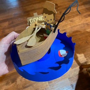 Puede incluir: Una figurita de papel de una persona en un bote con una ca&ntilde;a de pescar y un cartel que dice "Gone Fishin". El bote est&aacute; encima de una ola de papel azul con un flotador de pesca rojo y blanco.