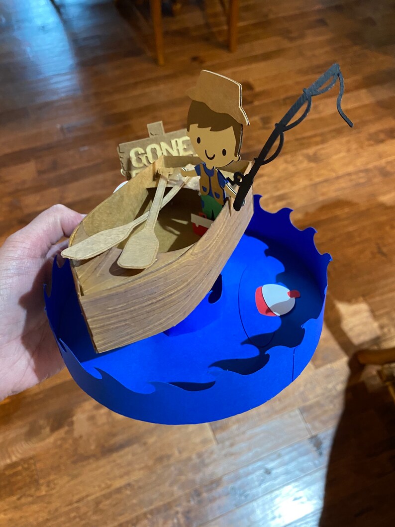 Puede incluir: Una figurita de papel de una persona en un bote con una ca&ntilde;a de pescar y un cartel que dice "Gone Fishin". El bote est&aacute; encima de una ola de papel azul con un flotador de pesca rojo y blanco.
