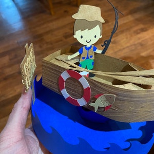 Puede incluir: Una manualidad de papel de un ni&ntilde;o con un sombrero y un chaleco marrones pescando desde un barco marr&oacute;n con un salvavidas. El barco est&aacute; sobre un fondo de olas azules. El cartel dice "Cast A Line".