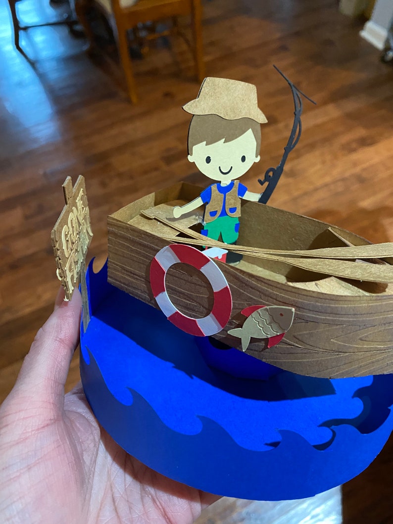 Puede incluir: Una manualidad de papel de un ni&ntilde;o con un sombrero y un chaleco marrones pescando desde un barco marr&oacute;n con un salvavidas. El barco est&aacute; sobre un fondo de olas azules. El cartel dice "Cast A Line".