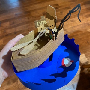Puede incluir: Una manualidad de papel de una persona en un bote con una ca&ntilde;a de pescar. El bote es marr&oacute;n y tiene dos remos. La persona lleva un sombrero marr&oacute;n y una camisa azul. El bote est&aacute; encima de una ola azul con una boya roja. El letrero en el bote dice "Gone Fishin".