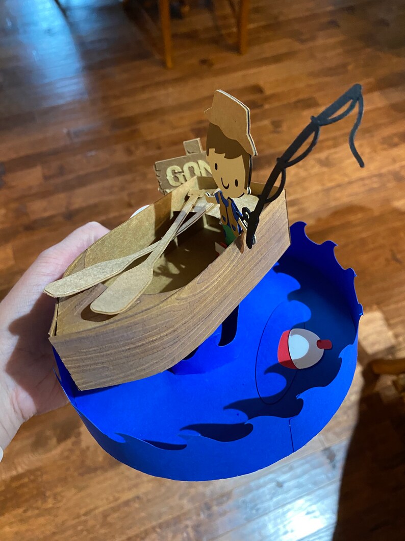 Puede incluir: Una manualidad de papel de una persona en un bote con una ca&ntilde;a de pescar. El bote es marr&oacute;n y tiene dos remos. La persona lleva un sombrero marr&oacute;n y una camisa azul. El bote est&aacute; encima de una ola azul con una boya roja. El letrero en el bote dice "Gone Fishin".