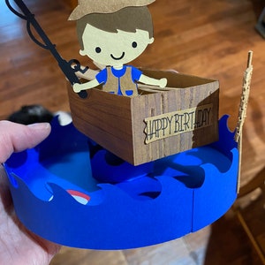 Puede incluir: Una tarjeta de cumplea&ntilde;os de papel con un pescador de dibujos animados en un bote con una ca&ntilde;a de pescar y las palabras "Feliz cumplea&ntilde;os" en el lateral. El bote est&aacute; sentado sobre olas azules.